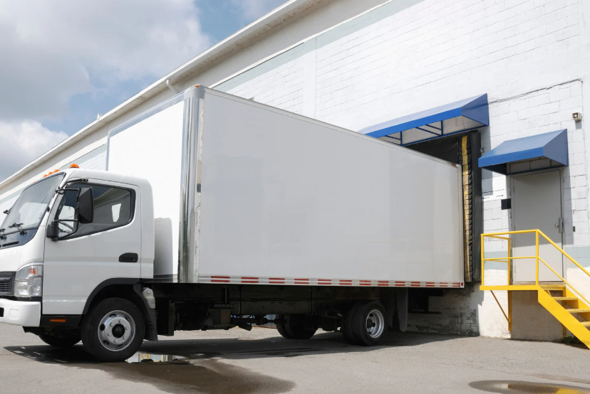 A white medium-duty box truck backed up to a commercial warehouse loading dock with yellow safety stairs, representing the type of delivery and commercial vehicles involved in serious truck accident injury cases in texas.