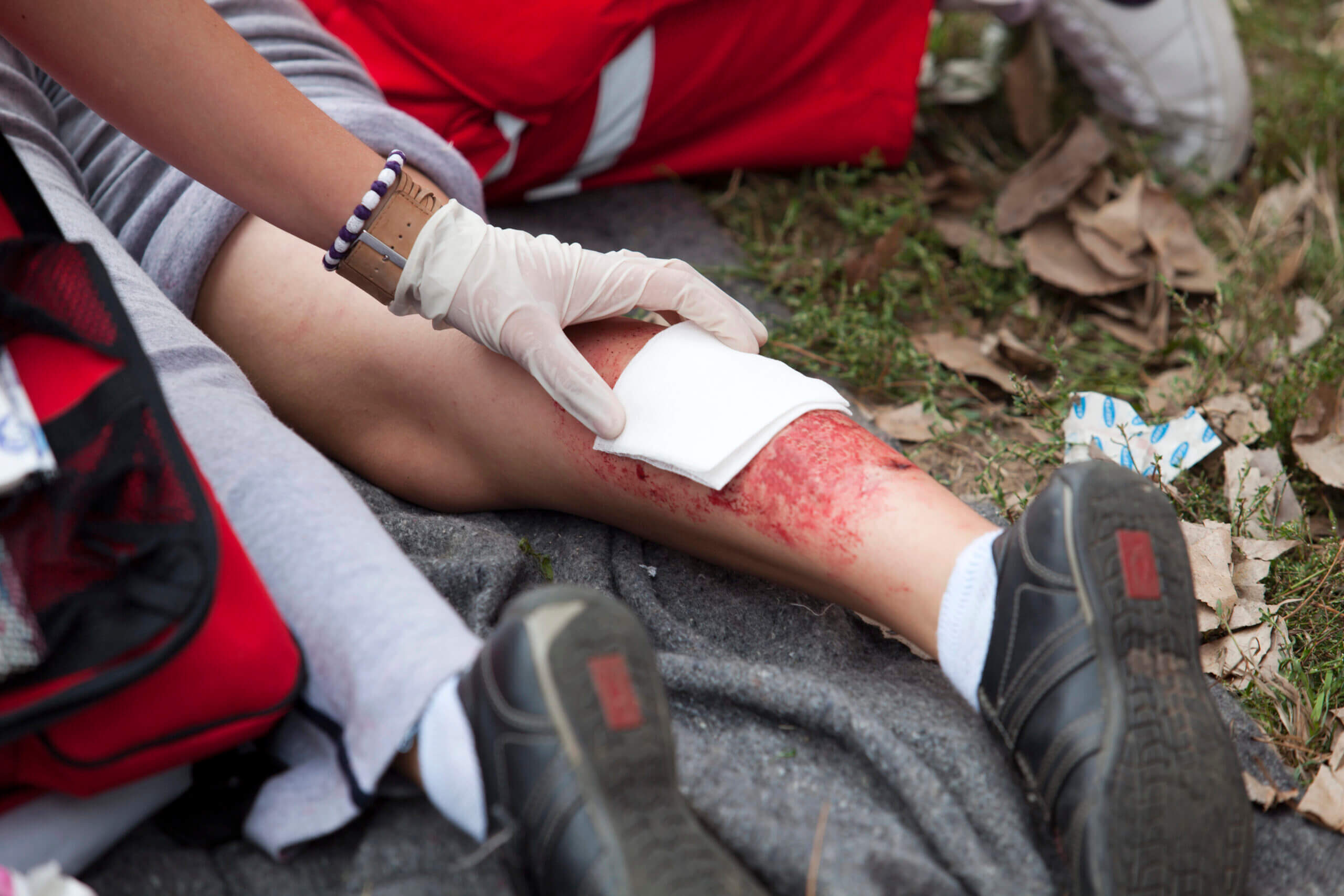 A first responder in a red uniform applying a gauze bandage to a severe road rash and laceration wound on a truck accident victim's leg at the crash scene.